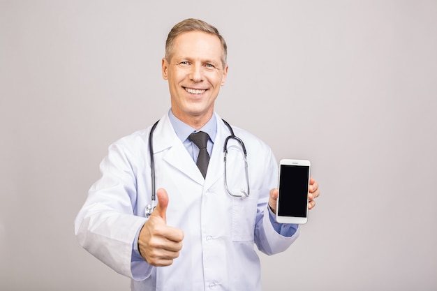 Hombre mayor feliz del doctor que muestra una pantalla en blanco del teléfono inteligente aislada en un fondo gris.