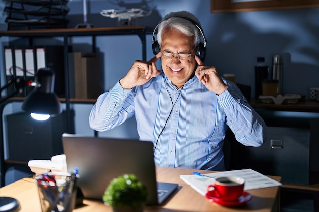 Hombre mayor hispano que usa auriculares de agente del centro de llamadas en la noche cubriendo las orejas con los dedos con expresión molesta por el ruido de la música a todo volumen concepto sordo