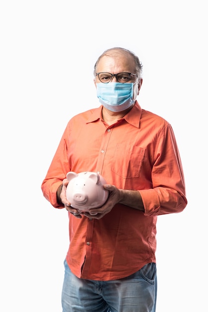 Hombre mayor jubilado indio que sostiene la hucha o la caja de dinero mientras usa la mascarilla en la pandemia