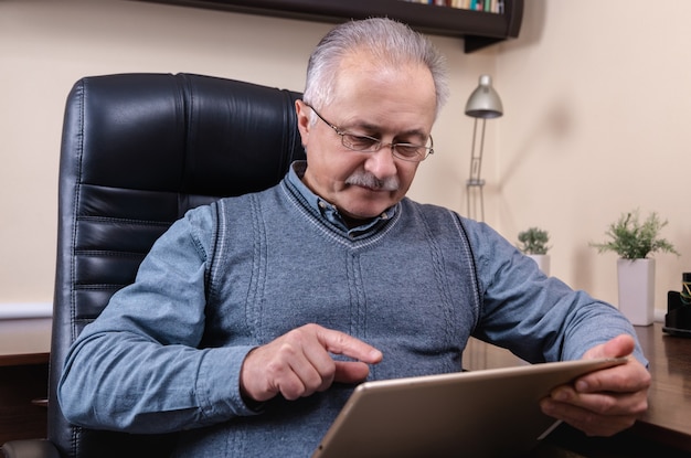 Hombre mayor leyendo noticias en tableta digital. Hombre mayor con tableta, sentado en el escritorio en casa. Tecnología moderna, concepto de comunicación