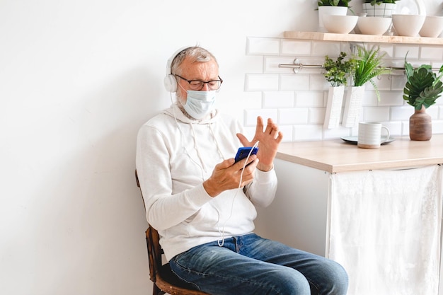 Hombre mayor con máscara médica en la cara usando la aplicación de teléfono inteligente para crear música de lista de reproducción Hombre mayor divirtiéndose con tecnología de teléfono móvil copia espacio Tecnología y alegre concepto de estilo de vida de los ancianos