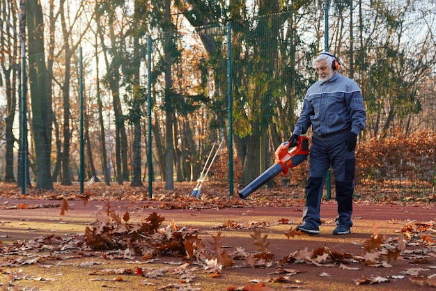 Hombre mayor con monos, auriculares y guantes limpiando el área vallada del parque de la ciudad de hojas secas de otoño