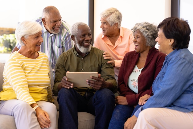 Hombre mayor multirracial sosteniendo una tableta digital mirando a sus amigos riéndose mientras se sienta en el sofá. Hogar de ancianos, tecnología inalámbrica, inalterado, unión, apoyo, vida asistida, jubilación.