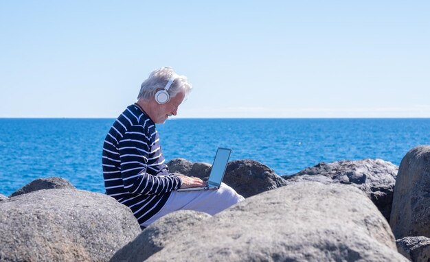 Hombre mayor de pelo blanco que trabaja en un portátil sentado al aire libre frente al mar con auriculares ancianos nómadas varones caucásicos disfrutando de la tecnología y las redes sociales