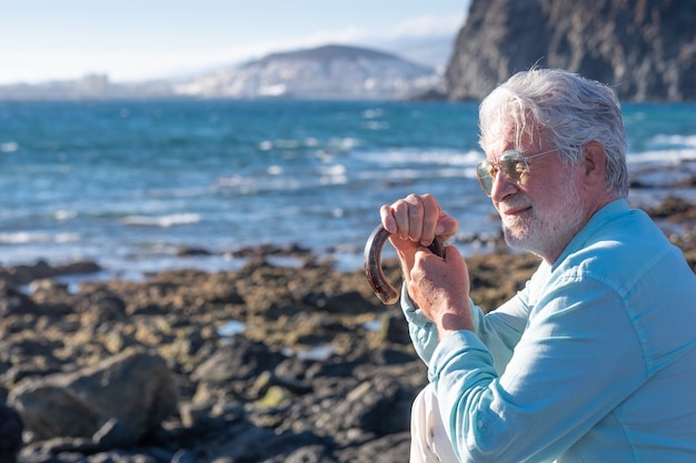 Hombre mayor, de pelo blanco, sosteniendo un bastón sentado en la playa mirando al horizonte sobre el agua