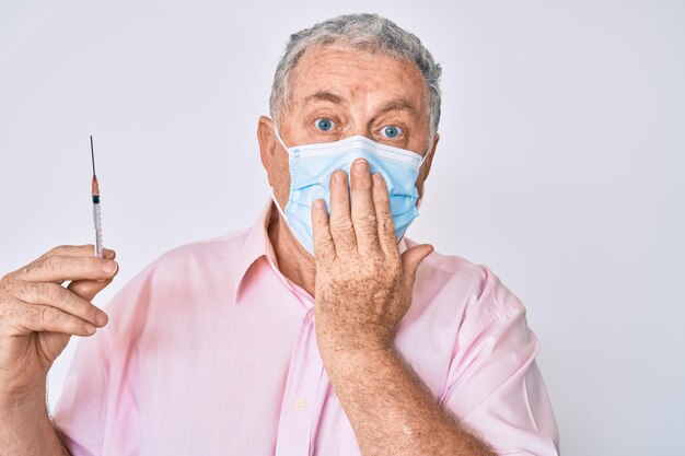Hombre mayor de pelo gris con máscara médica sosteniendo una jeringa que cubre la boca con la mano sorprendida y asustada por el error expresión sorprendida