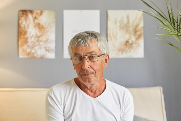 Un hombre mayor de pelo gris tranquilo con camiseta blanca informal y gafas sentado en el sofá de casa en el interior de la sala de estar mirando la cámara con expresión enfocada