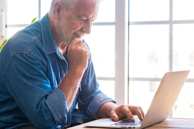 Hombre mayor pensativo, con barba blanca, sonríe mirando su computadora portátil navegando por la red. Gente moderna y nueva tecnología