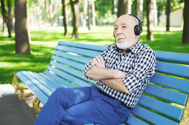 Hombre mayor pensativo escuchando música al aire libre. Anciano en auriculares sentado en el banco en el parque, espacio de copia