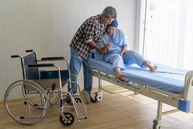 Un hombre mayor que ayuda a una mujer paciente con cáncer que usa un pañuelo en la cabeza que se traslada a una silla de ruedas en el hospital