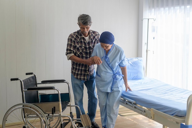 Un hombre mayor que ayuda a una mujer paciente con cáncer que usa un pañuelo en la cabeza que se traslada a una silla de ruedas en el hospital