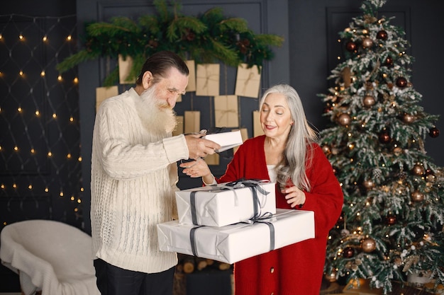 Hombre mayor que presenta un regalo de navidad a su esposa de pie cerca del árbol de navidad