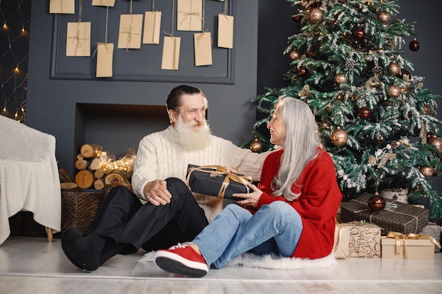 Hombre mayor que presenta un regalo de navidad a su esposa sentada cerca del árbol de navidad