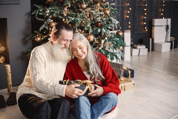 Hombre mayor que presenta un regalo de navidad a su esposa sentada cerca del árbol de navidad