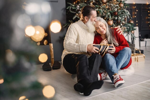 Hombre mayor que presenta un regalo de navidad a su esposa sentada cerca del árbol de navidad