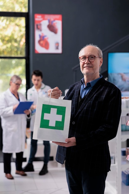 Hombre mayor que sostiene el bolso de compras de la compra en retrato de la farmacia. Farmacia anciano cliente sonriente comprando medicamentos recetados, parado en boticario y mirando a la cámara