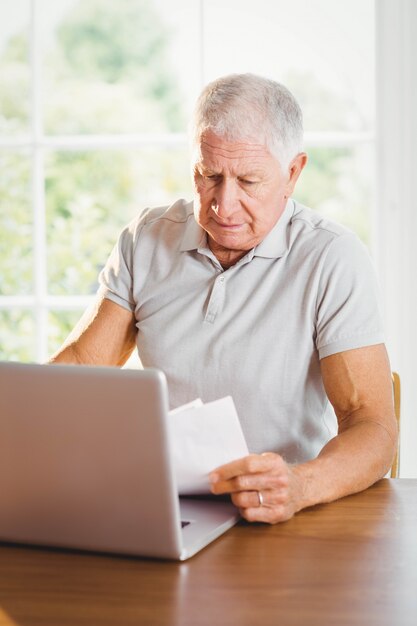 Hombre mayor que sostiene las hojas y que usa la computadora portátil en casa