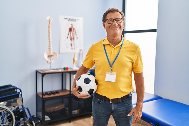 Hombre mayor que trabaja en la clínica de fisioterapia deportiva con aspecto positivo y feliz de pie y sonriendo con una sonrisa segura mostrando los dientes