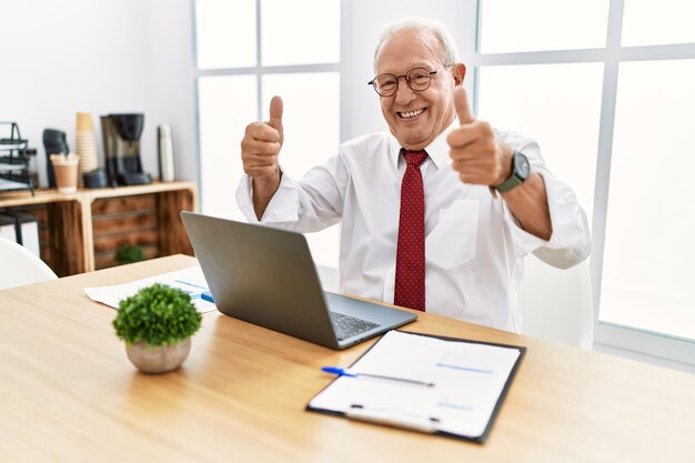 Hombre mayor que trabaja en la oficina usando una computadora portátil que aprueba hacer un gesto positivo con la mano, pulgares arriba sonriendo y feliz por el éxito. gesto ganador.