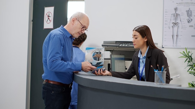 Hombre mayor que usa tarjeta de crédito para pagar la consulta en el mostrador de registro con recepcionista asiática en el vestíbulo. Hacer el pago de transacciones electrónicas para la visita de control y la cita, pagar el tratamiento.