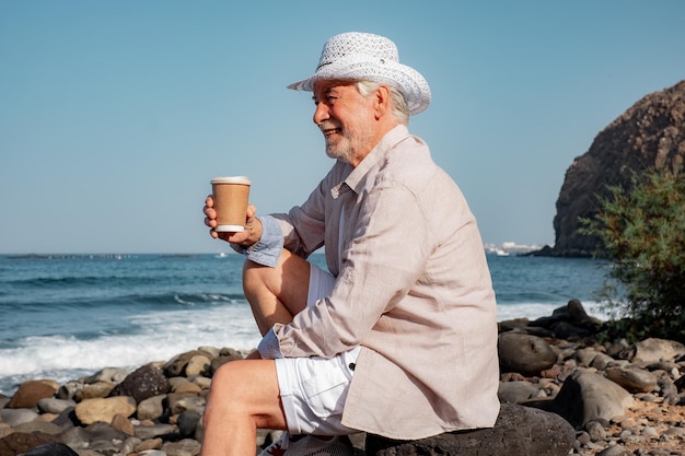 Hombre mayor relajado sentado en la playa sosteniendo una taza de café mirando hacia la jubilación de vacaciones