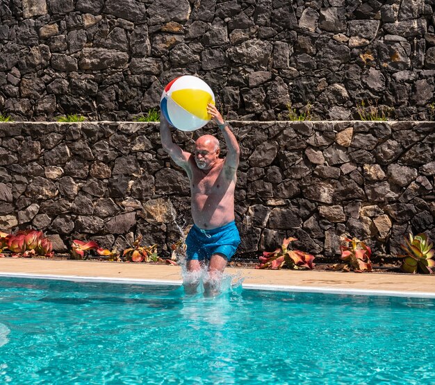 Hombre mayor salta a la piscina con un gran globo en las manos. Verano y diversión. Jugando con amigos. Barba y pelos blancos