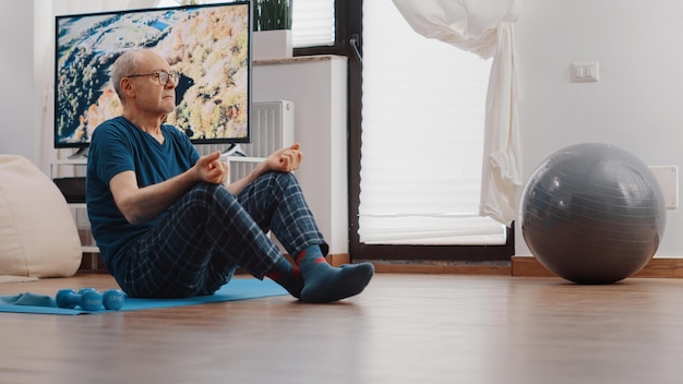 Hombre mayor sentado en posición de loto y haciendo meditación en estera de yoga. Jubilado Zen respirando para meditar y relajarse después del ejercicio. Anciano meditando para el bienestar y la recreación