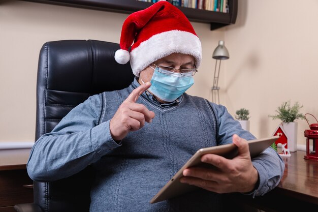 Hombre mayor con sombrero de santa claus habla con dispositivo de tableta para videollamadas amigos y niños. La habitación está decorada de forma festiva. Navidad durante el coronavirus.