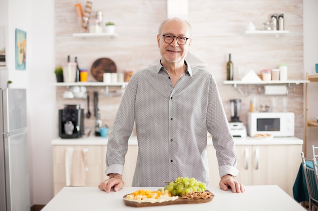 Hombre mayor sonriente en la cocina de casa mirando a la cámara con variedad de sabroso queso y uvas en la mesa.