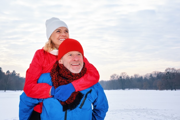 Hombre mayor sonriente dando a mujer madura a cuestas en el parque de invierno cubierto de nieve