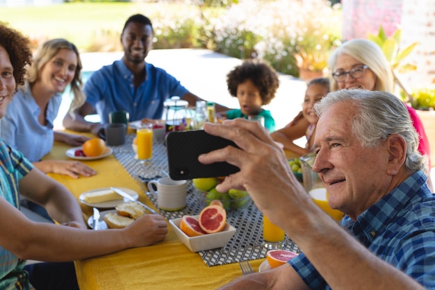 Un hombre mayor sonriente tomándose un selfie a través de un teléfono inteligente con una familia multirracial mientras desayunaba