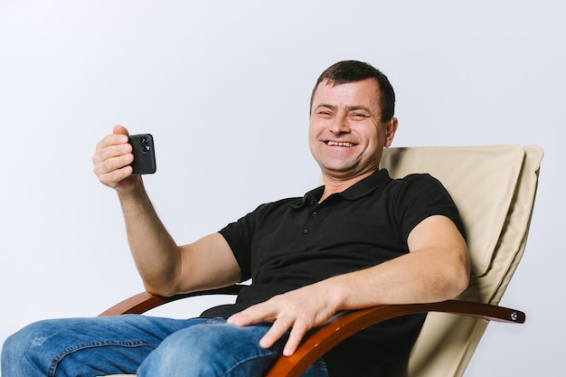 Hombre mayor sordo sonriente con un dispositivo auditivo en un sillón de cuero, se comunica en un teléfono inteligente