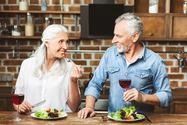 Hombre mayor sosteniendo una copa de vino tinto mirando a su esposa comiendo ensalada en la cocina