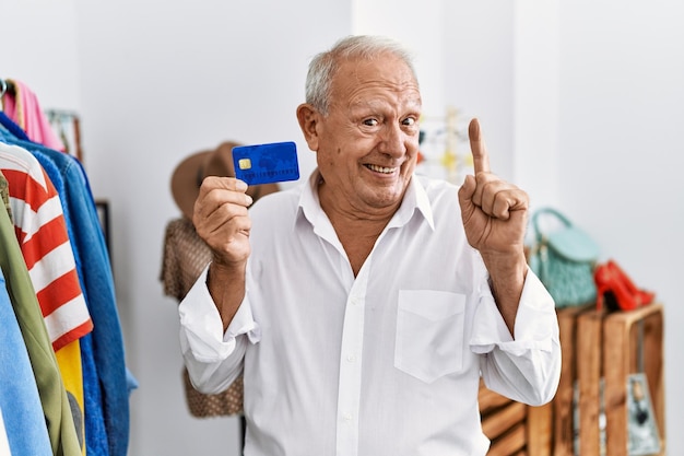 Hombre mayor en la tienda minorista con tarjeta de crédito sonriendo con una idea o pregunta señalando con el dedo con la cara feliz número uno