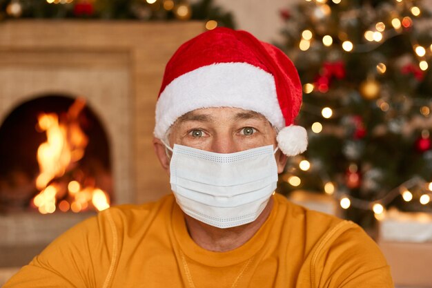 Hombre mayor vestido con una máscara protectora en su rostro y sombrero de Navidad mira a la cámara, posando en la sala festiva con chimenea borrosa y árbol de Navidad. Quédate en casa.