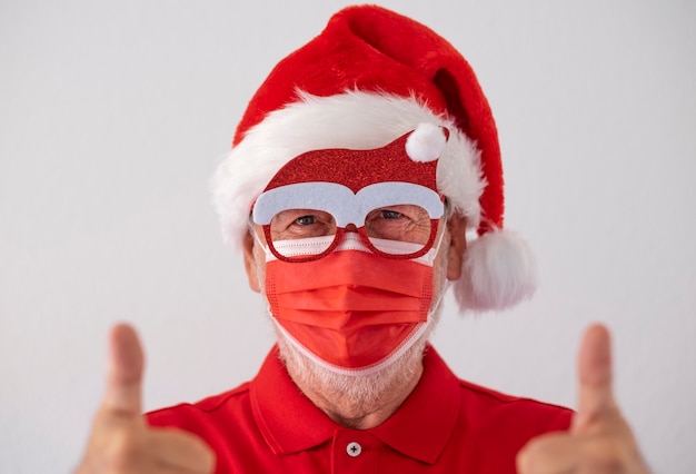 Foto un hombre mayor vestido de rojo como papá noel con una mascarilla quirúrgica debido al coronavirus y hace una señal de optimismo. feliz navidad en la época del virus covid-19.