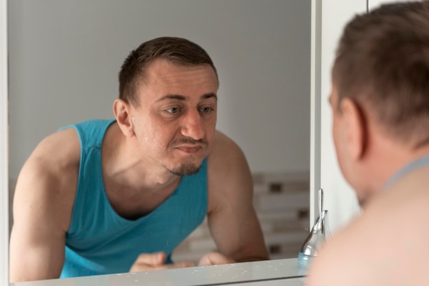 Hombre de mediana edad sin afeitarse la cara se inclina sobre el fregadero y mira con desagrado el reflejo en el espejo Higiene matutina