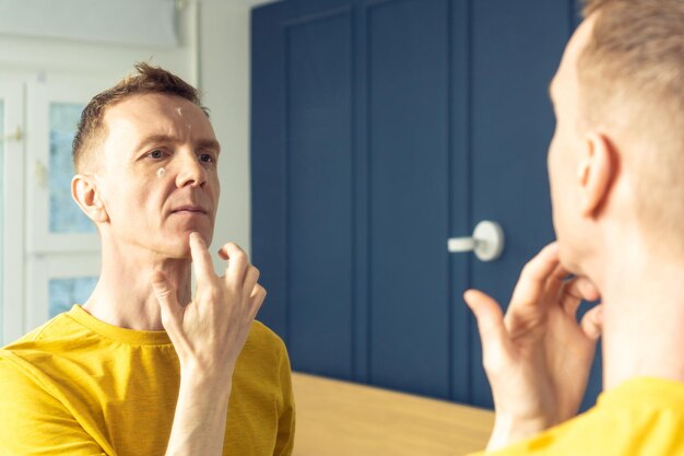 Un hombre de mediana edad aplica crema hidratante en áreas faciales claras Retrato masculino en el espejo de la habitación Rutina matutina de cuidado de la piel