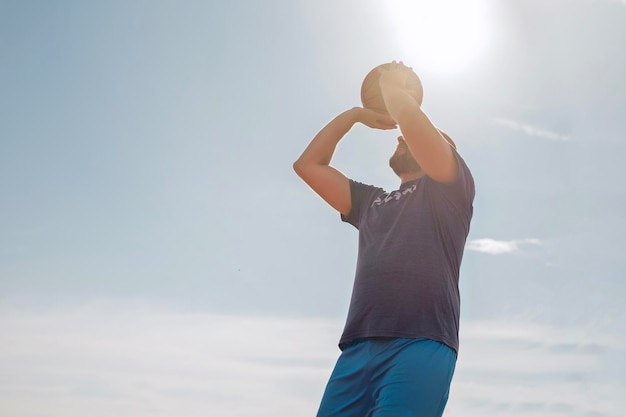 Un hombre de mediana edad apunta a lanzar una pelota de baloncesto a la canasta. Un hombre haciendo deporte contra el fondo de un cielo azul bajo los rayos del sol poniente.