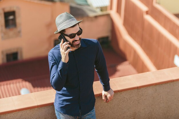 Hombre de mediana edad atractivo con sombrero y gafas de sol hablando con un teléfono inteligente de pie en el balcón.