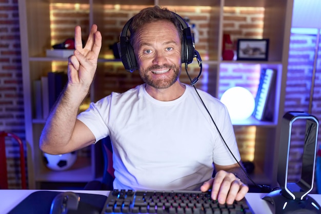 Hombre de mediana edad con barba jugando videojuegos con auriculares que aparecen y señalan con los dedos número tres mientras sonríe confiado y feliz