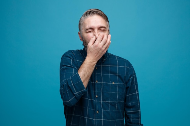 Hombre de mediana edad con cabello gris en camisa de color oscuro cerrando la nariz con los dedos que sufren de hedor de pie sobre fondo azul.