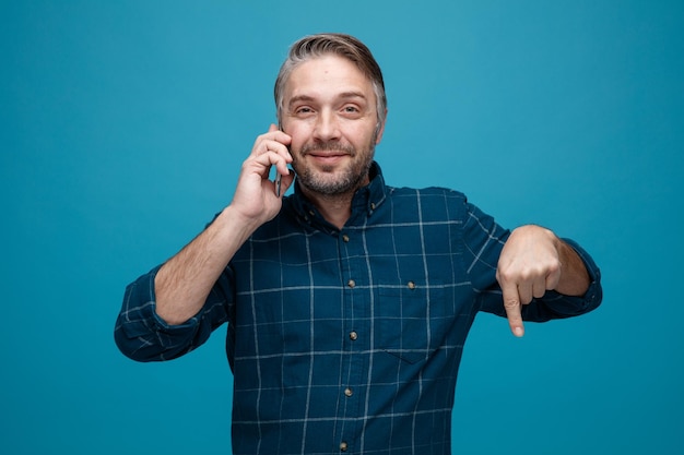 Hombre de mediana edad con cabello gris en camisa de color oscuro hablando por teléfono móvil sonriendo señalando con el dedo índice hacia abajo de pie sobre fondo azul.