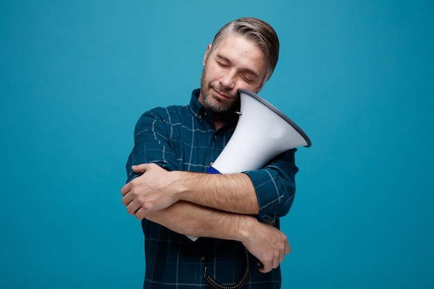 Foto hombre de mediana edad con cabello gris en camisa de color oscuro con megáfono luciendo feliz y complacido de pie sobre fondo azul.