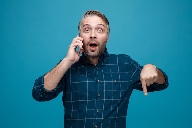 Hombre de mediana edad con cabello gris en camisa de color oscuro mirando a la cámara asombrado y sorprendido mientras habla por teléfono móvil apuntando con el dedo índice hacia abajo de pie sobre fondo azul