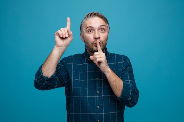 Hombre de mediana edad con cabello gris en camisa de color oscuro mirando a la cámara con cara seria haciendo gesto de silencio con el dedo en los labios apuntando con el dedo índice hacia arriba de pie sobre fondo azul