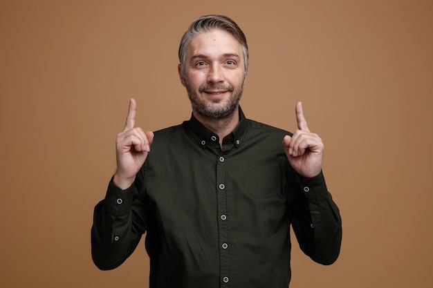 Hombre de mediana edad con cabello gris en camisa de color oscuro mirando a la cámara con una sonrisa en la cara feliz apuntando con los dedos índices de pie sobre fondo marrón