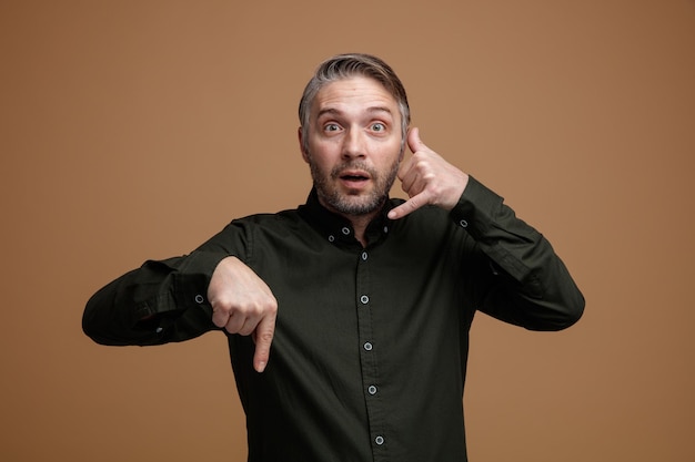 Hombre de mediana edad con cabello gris en camisa de color oscuro mirando a la cámara sorprendido haciendo un gesto de llamarme apuntando con el dedo índice hacia abajo de pie sobre fondo marrón