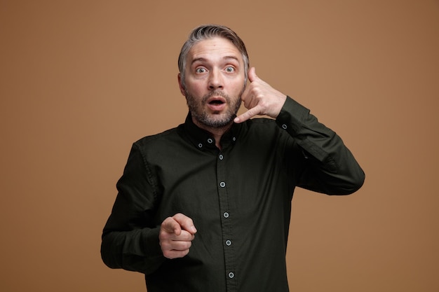 Hombre de mediana edad con cabello gris en camisa de color oscuro mirando a la cámara sorprendido haciendo un gesto de llamarme con la mano apuntando con el dedo índice a la cámara de pie sobre fondo marrón