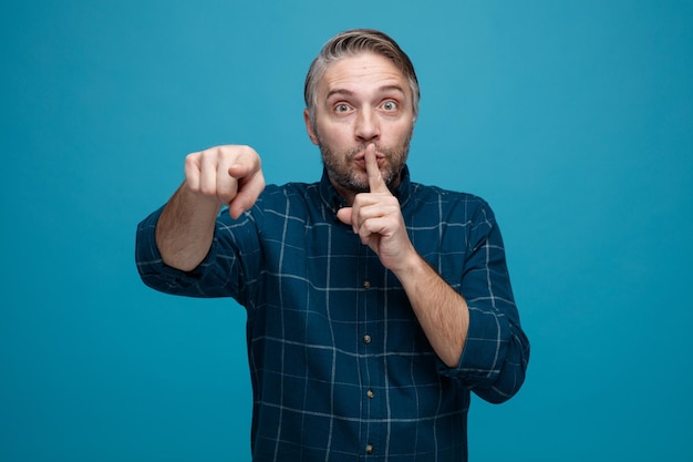 Hombre de mediana edad con cabello gris en camisa de color oscuro mirando a la cámara sorprendido haciendo gesto de silencio con el dedo en los labios apuntando con el dedo índice a la cámara de pie sobre fondo azul